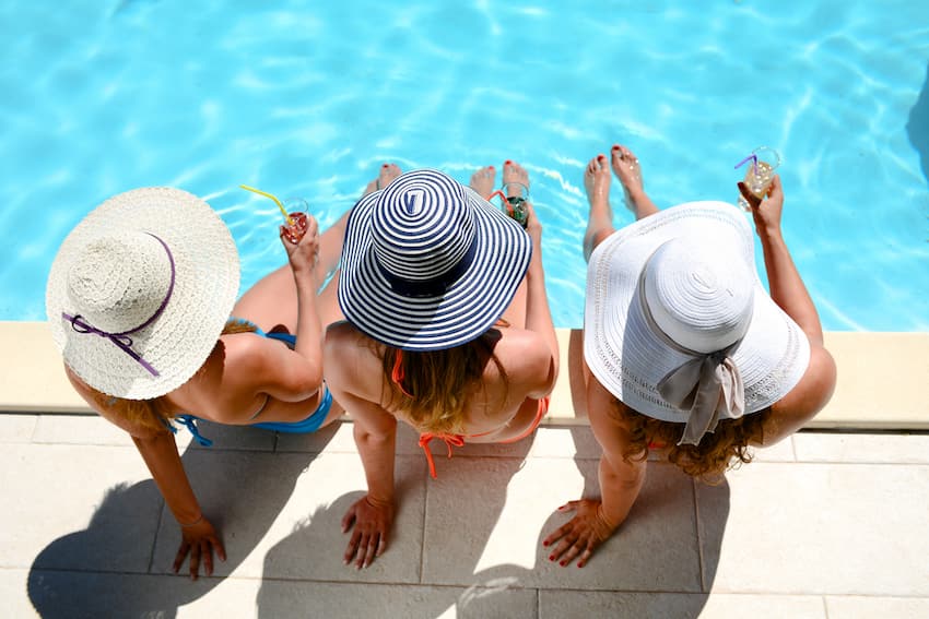 three friends with stylish hats sit by the pool with their feet in the water