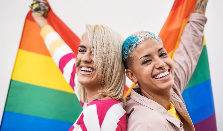 Two people painted in pride flag colors hold a rainbow flag and smile