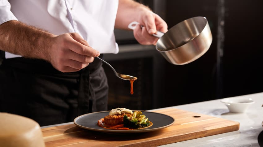 a cook drizzles juice onto a plate with a small portion of food on it